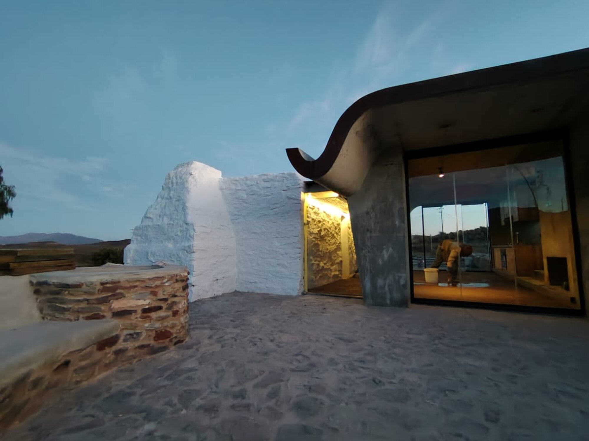Sheep Herder's Cottage at dusk — lime-washed stone wall, the concrete shell roof and glass, the warm interior beyond